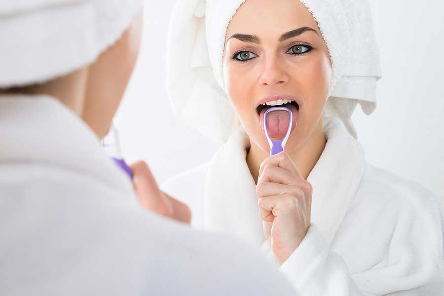 A woman using a tongue scraper to brush her tongue in the mirror.